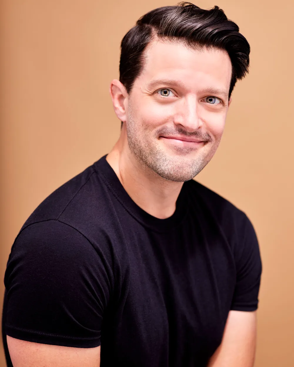 A headshot of Pacek, in a black shirt smiling against a soft pink colored backdrop