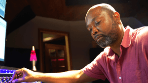 Coles, a Black man, sits at an illuminated, colorful soundboard in a dim studio