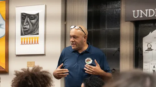 Close-up on Still, a bald Black man wearing jeans and a navy blazer, over the heads of people listening to him.