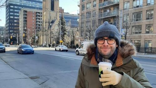 Samuel, a white man, stands smiling on a city street wearing a hat, gloves, and parka. He holds a cup of coffee.