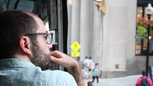 Samuel, a white man seen in profile from over his shoulder, looks thoughtfully out on a city street.