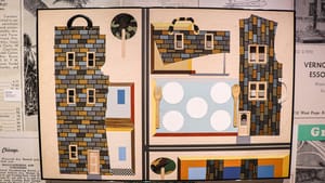 A collage-style piece in tan, orange, & blue, including brick buildings that have suitcase handles, and a wood table setting