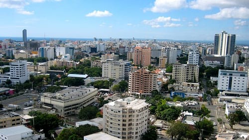 Arial photo of square and rectangular tan, white and brown buildings in a cityscape under a bright blue sky with a few clouds