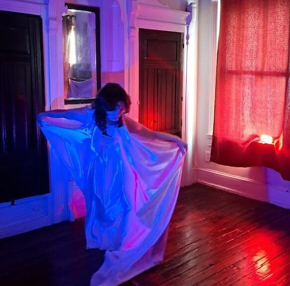 A person in a silky white garment with a long train sweeps it around them, lit by dramatic blue and pink light.
