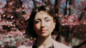 Sara Mae in a light jacket and black top with white paper floral spirals over their chest. The photo is fuzzy, out of focus