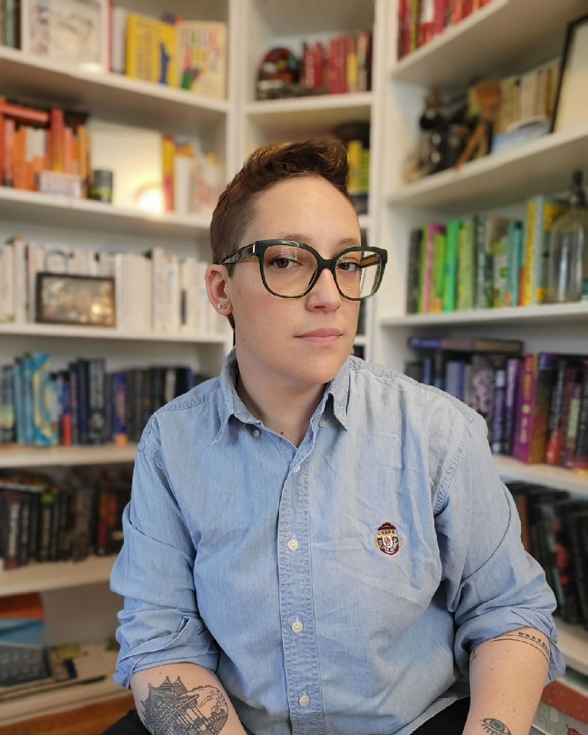 Profile of Novic. They are wearing glasses and a button down shirt, tattoos on their arm, books on shelves in the background