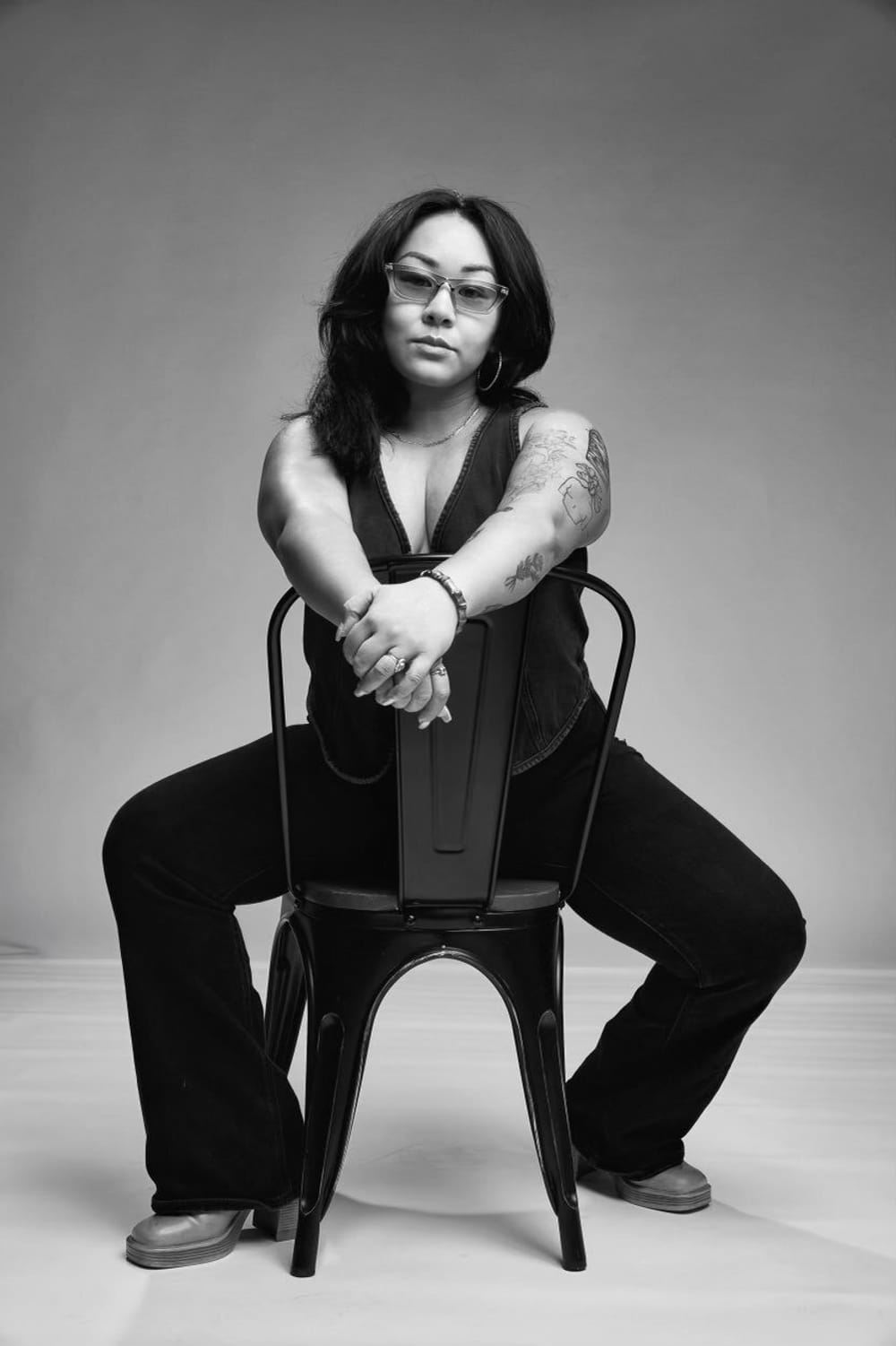B&W portrait of Porkalob, a Filipino woman (she/they) in all black, tattoos on her arms as she sits on a reversed black chair