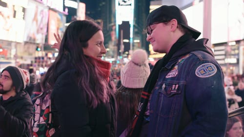 Riley and Sav stand facing each other, seen in profile at night in Times Square. They smile lovingly at each other.