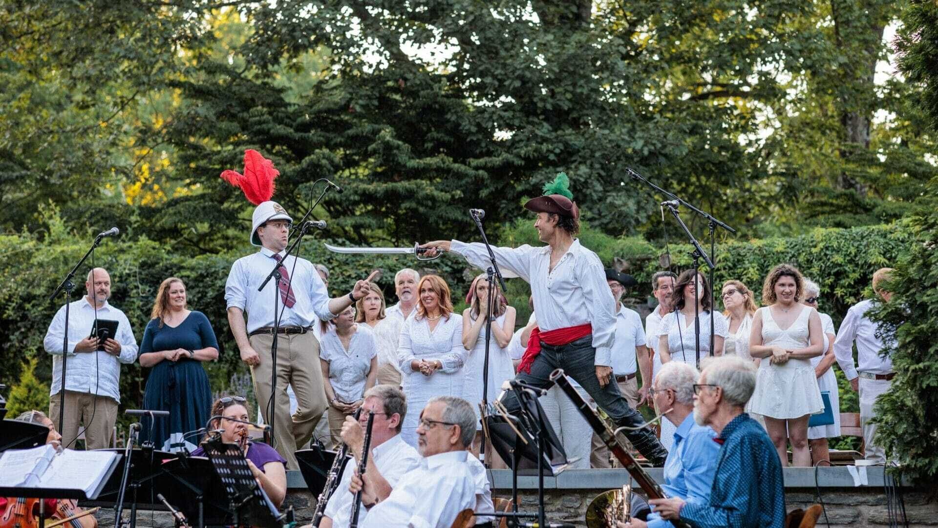 Two performers, outdoor stage; one in casual, other in a pirate outfit w/ a sword. An orchestra in the pit in front of stage