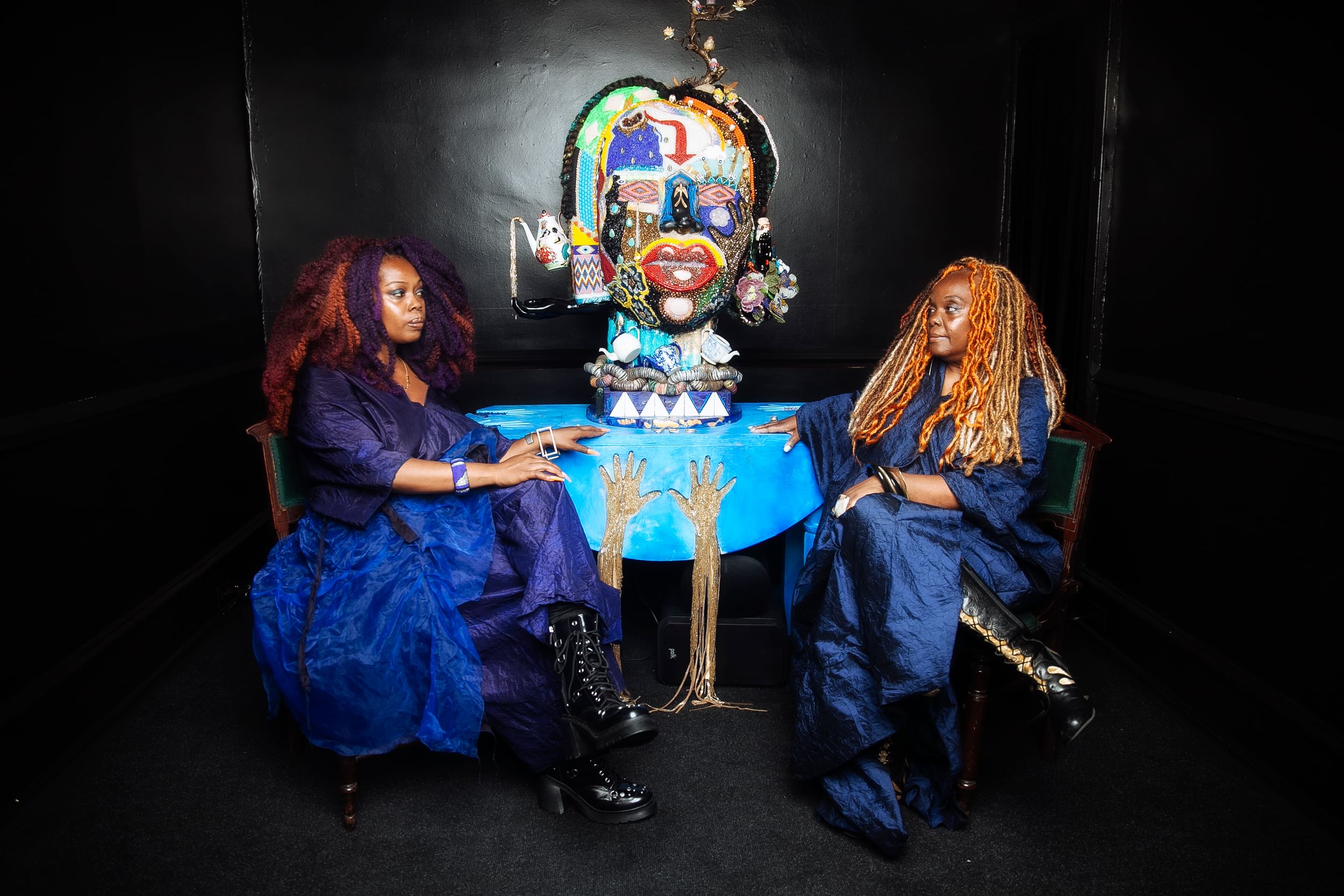 The DuBois women, in colorful bronze & purple coifs, sit by the artwork with serious expressions, wearing loose blue dresses