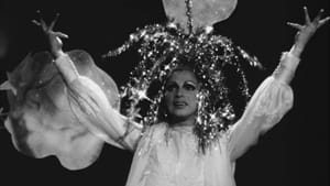 Film still. A performer in white, a sparkling floral-looking headdress, against a black backdrop