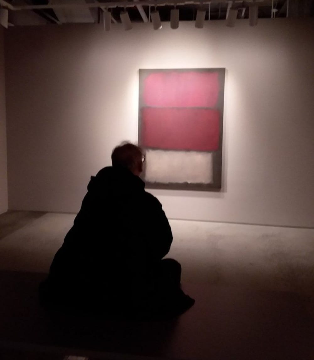 A shadowy, silhouetted image seen from behind a man regarding a red and white Rothko painting in a gallery.