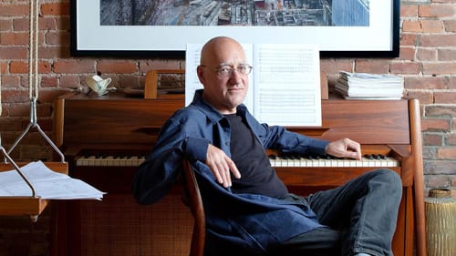 Currier, a bald white man with glasses, wearing denim, sits comfortably sideways to a piano, left hand poised on the keys.