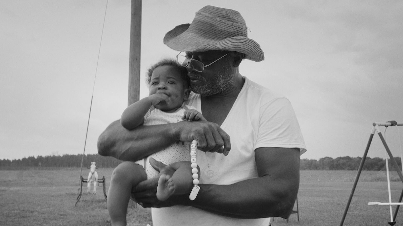 An elder Black man holds a baby, close up in the shot, a wide, open area with trees lining in the distance. B&W image.