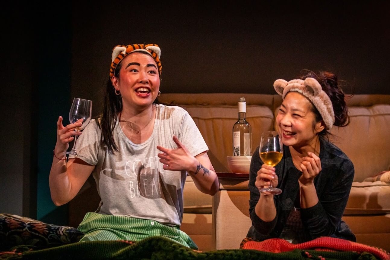 Li and Ngo, in cozy clothes and plush cat-ear headbands, laugh together in front of a couch, holding wineglasses.