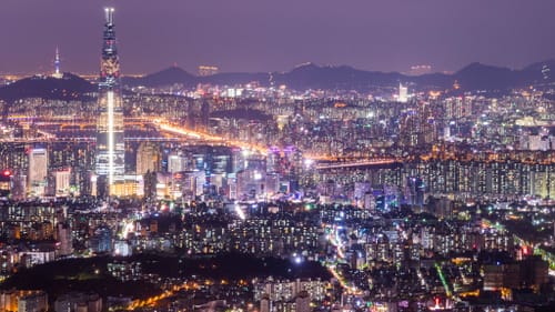 A nighttime photo of Seoul, misty purple sky and mountains beyond thousands of buildings spangled with light.