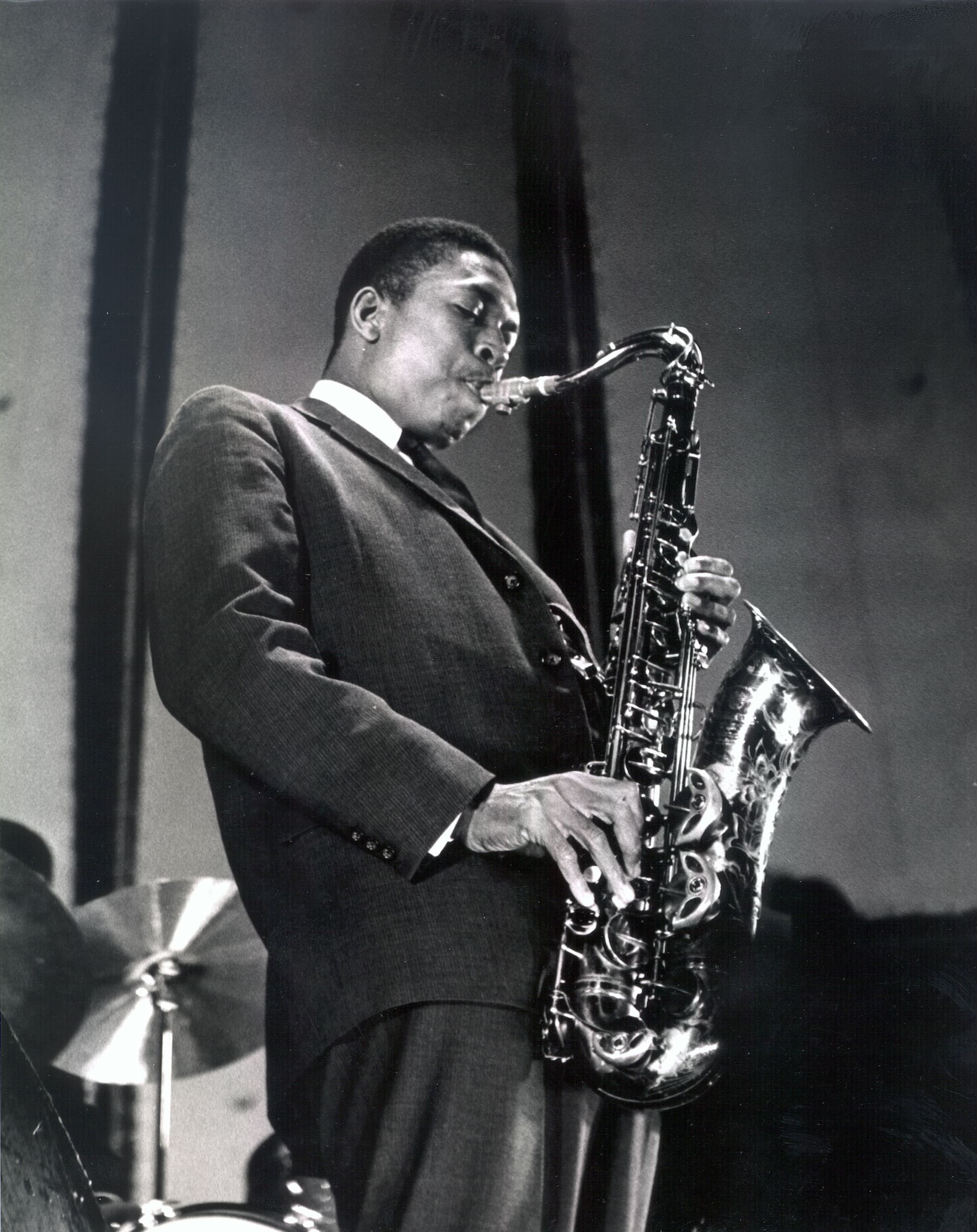 Vintage black & white photo of Coltrane playing a shiny sax while wearing a dark suit.