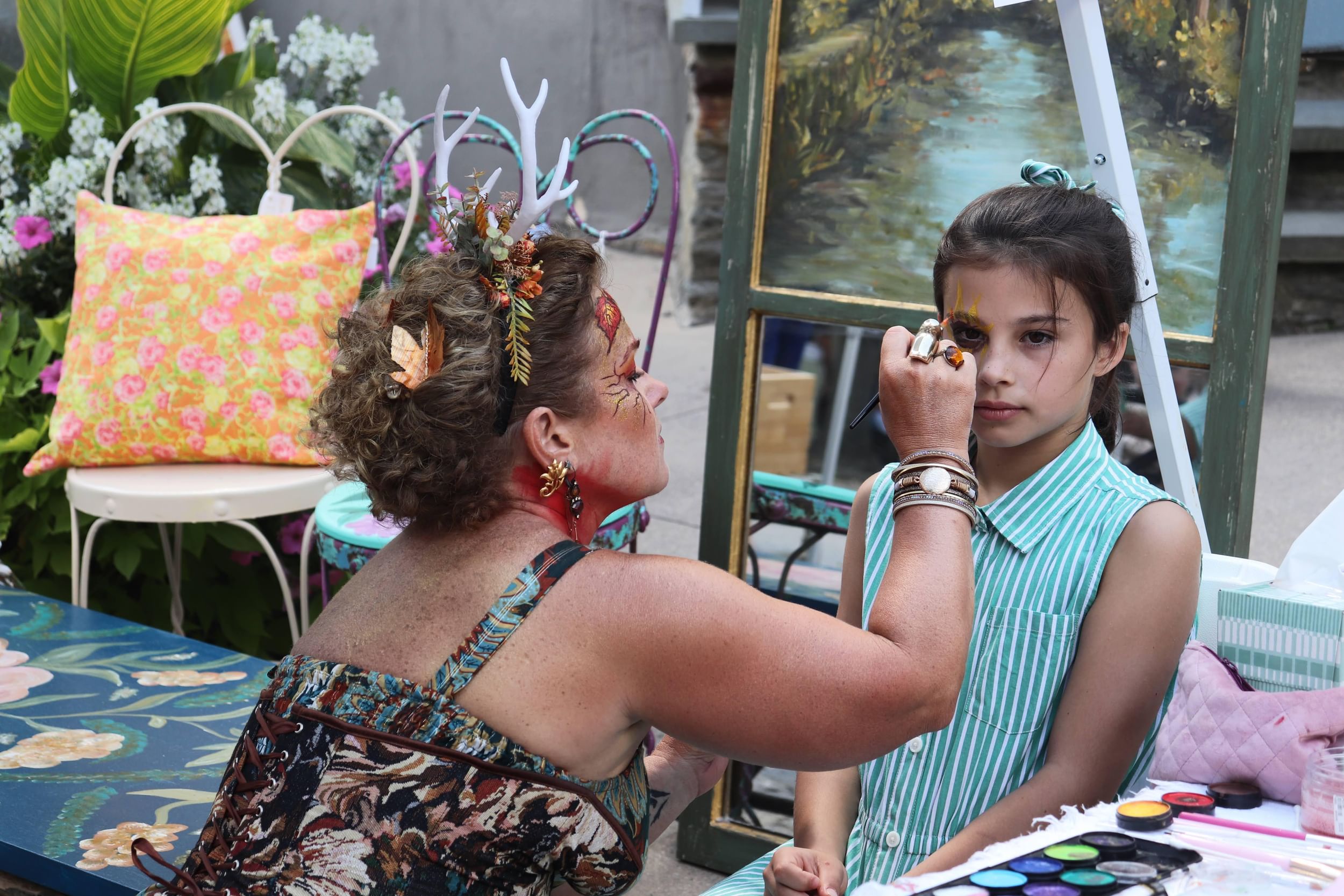 A girl with brown hair and a striped turquoise dress sits still while a fancifully decorated artist paints her face.