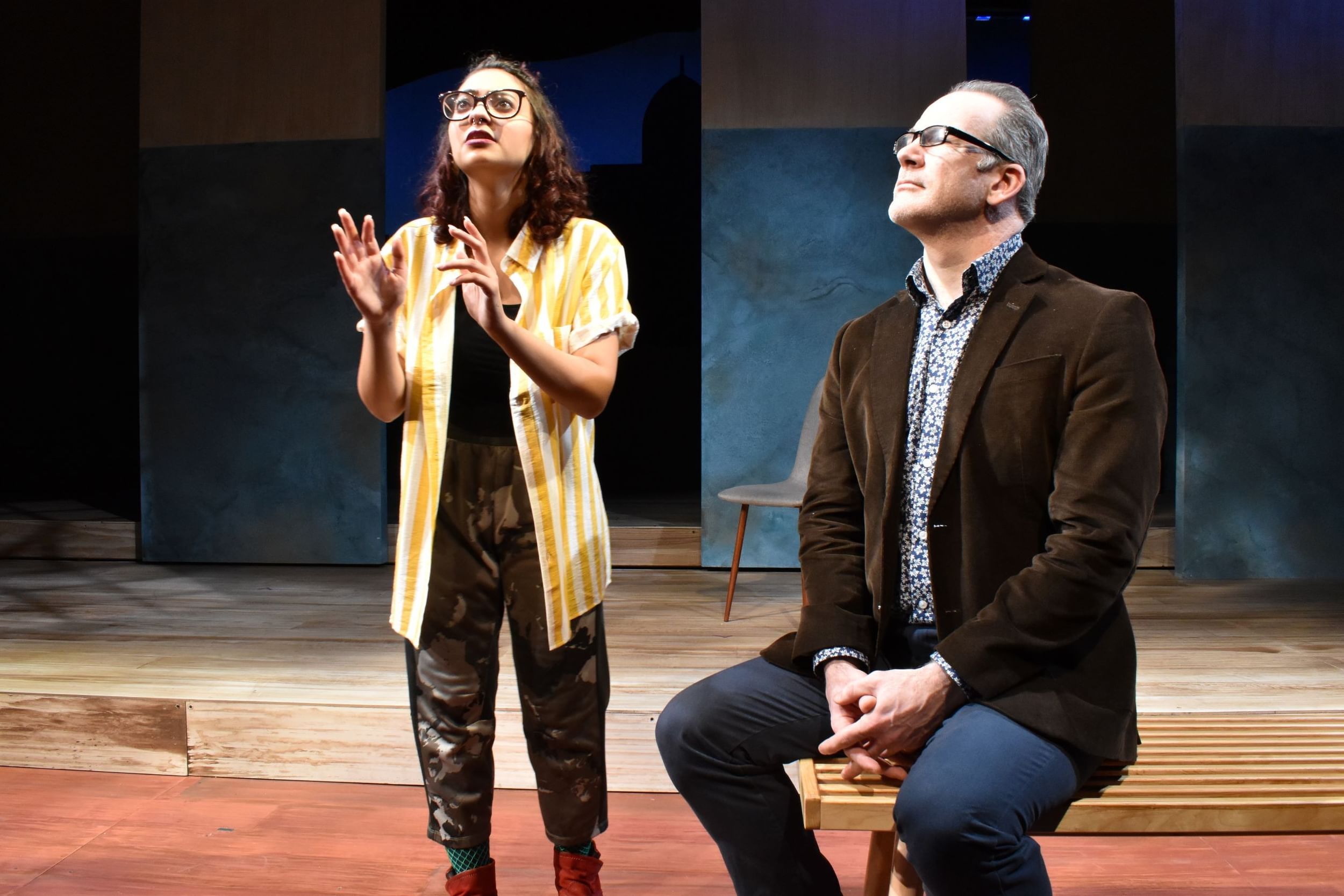 Scene from Settlements. Khalil, in a yellow-striped shirt, talks to Rishard, sitting on a bench, paying attention to Khalil.