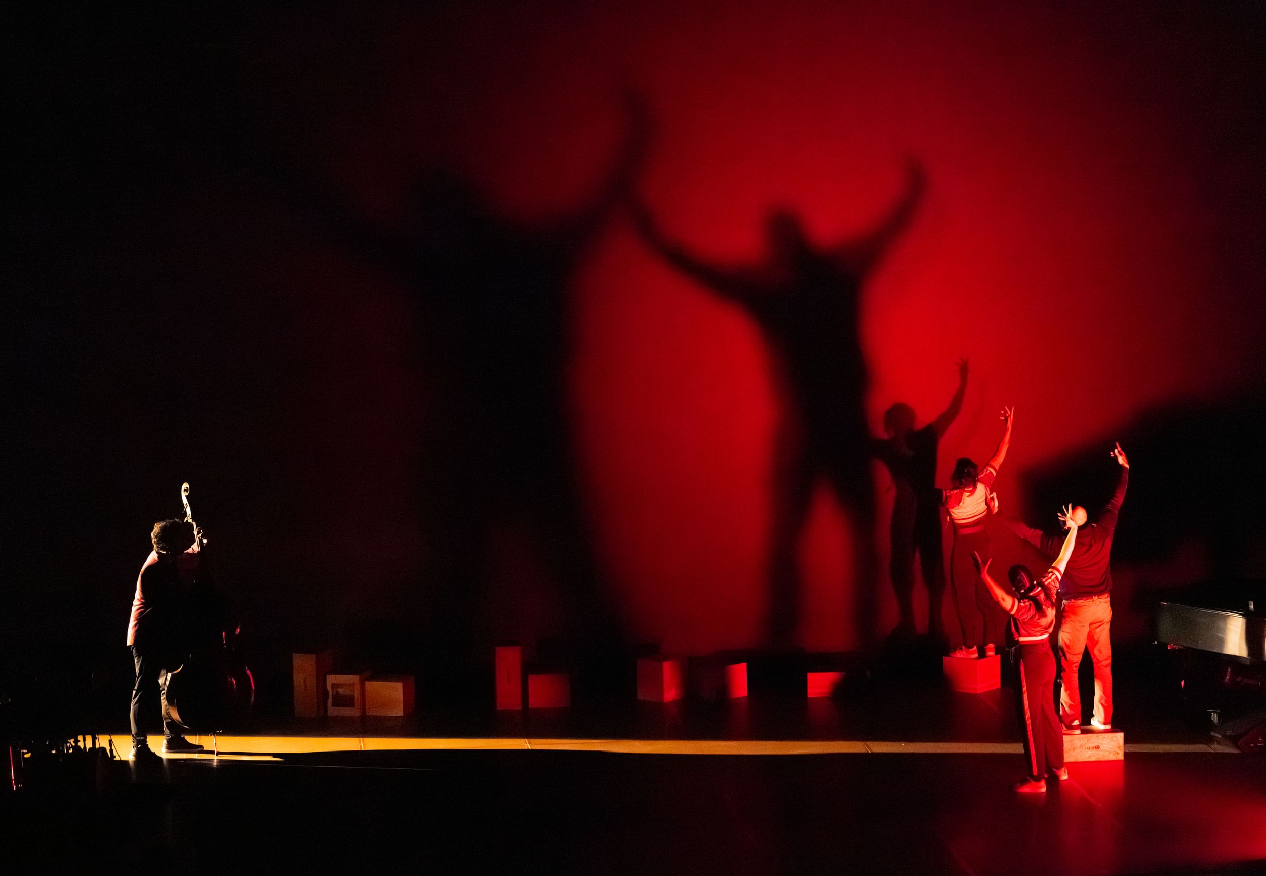 On a murky red-lit stage, three dancers at right with arms extended cast large, dramatic shadows on the wall behind them.
