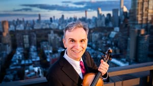 On a city rooftop, Shaham, a white man with gray hair, smiles and holds his violin, the blurry gray cityscape behind him.