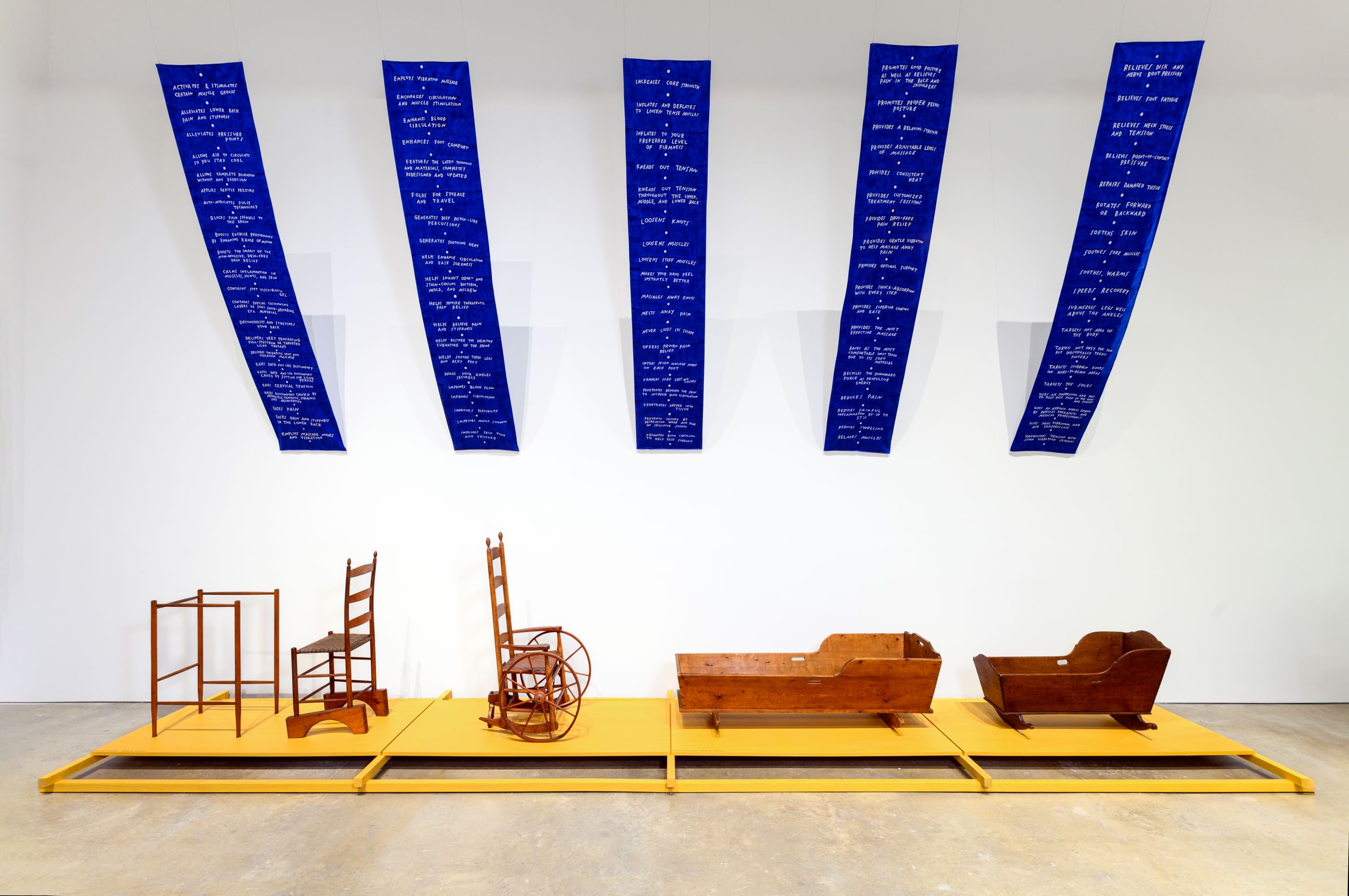 5 pieces of wooden Shaker furniture, including a wheelchair and two cradles, lined up in a white gallery below blue banners