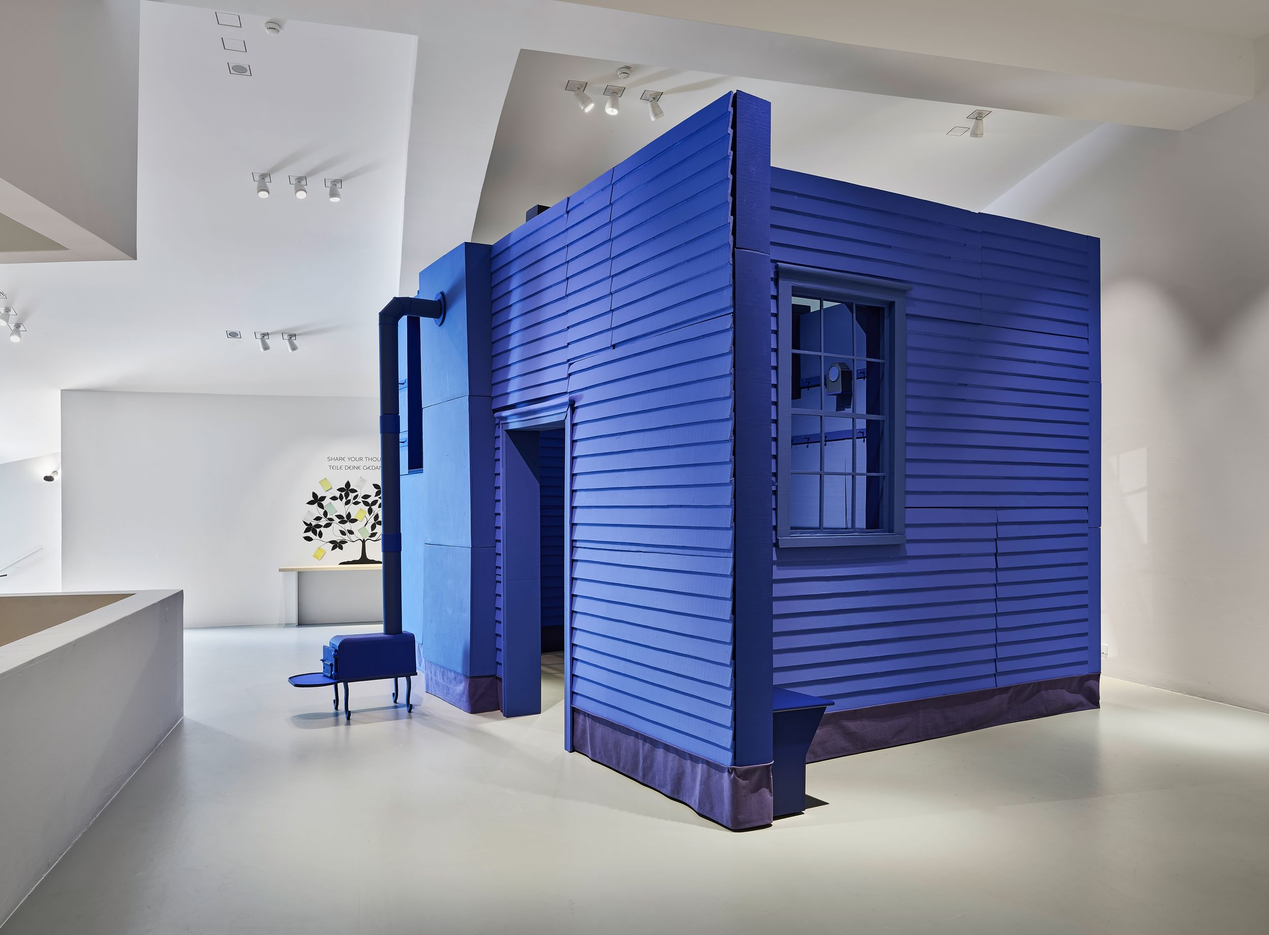 Large, striking, bright-blue structure with a door, siding, stovepipe, and windows in the spare white gallery at ICA.