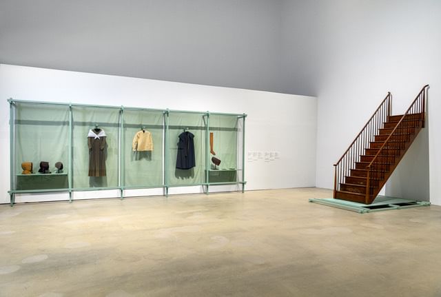 Historic Shaker clothing items on simple hangers, next to a fine wooden staircase attached to the white gallery wall.