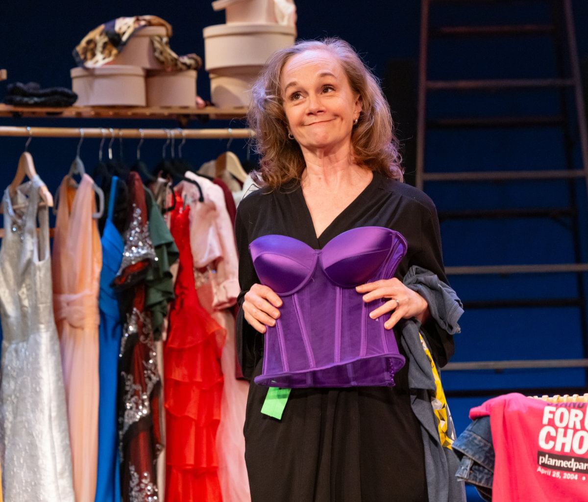 Childs, a 57-year-old white woman in a black dress, smiles ruefully as she holds up a purple satin corset.