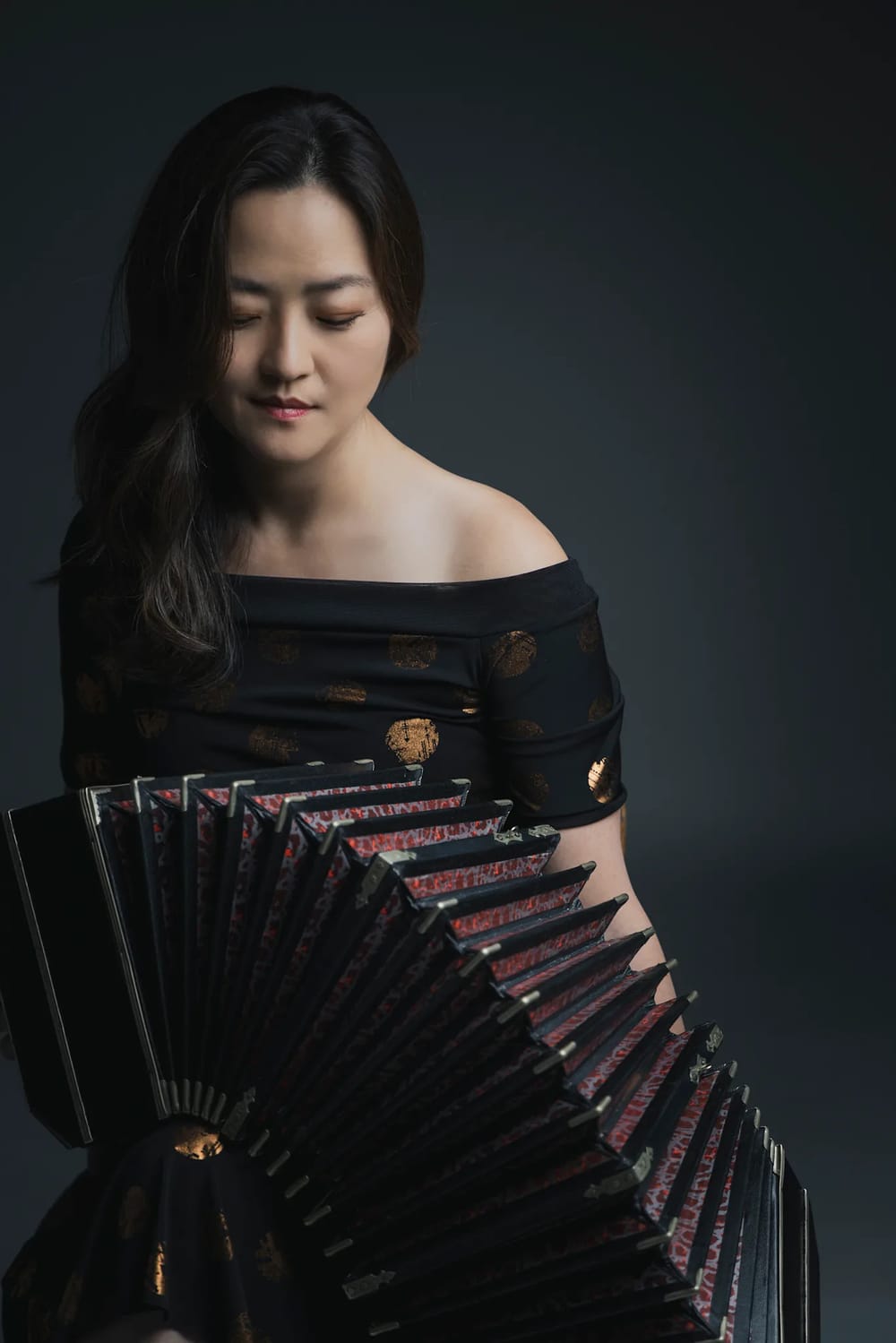 Cho, a Korean woman, looks down at her bandoneon, an accordion-like instrument. It’s extended out, falling over her lap