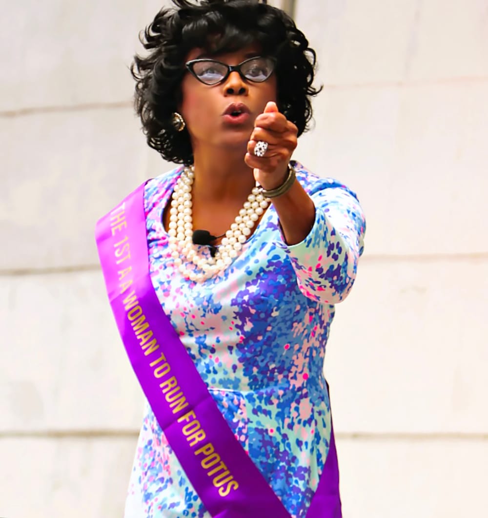 A Black woman in a purple/white dress with banner that reads “first AA woman to run for POTUS” motions at camera