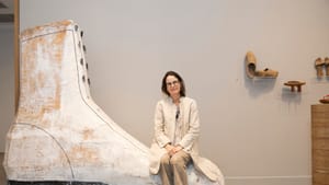 Kearney sits on a sculptured, large, mostly white boot several times her size. Other shoes rest on shelves in the gallery.