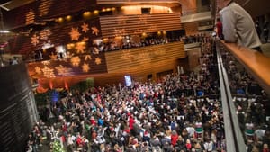 Wide shot of a large audience fills the indoors of Kimmel Center, with seasonal lights cast over the walls