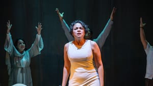 With chorus members raising arms behind, KO poses with an intent expression by a bed, in a sleeveless beige dress.