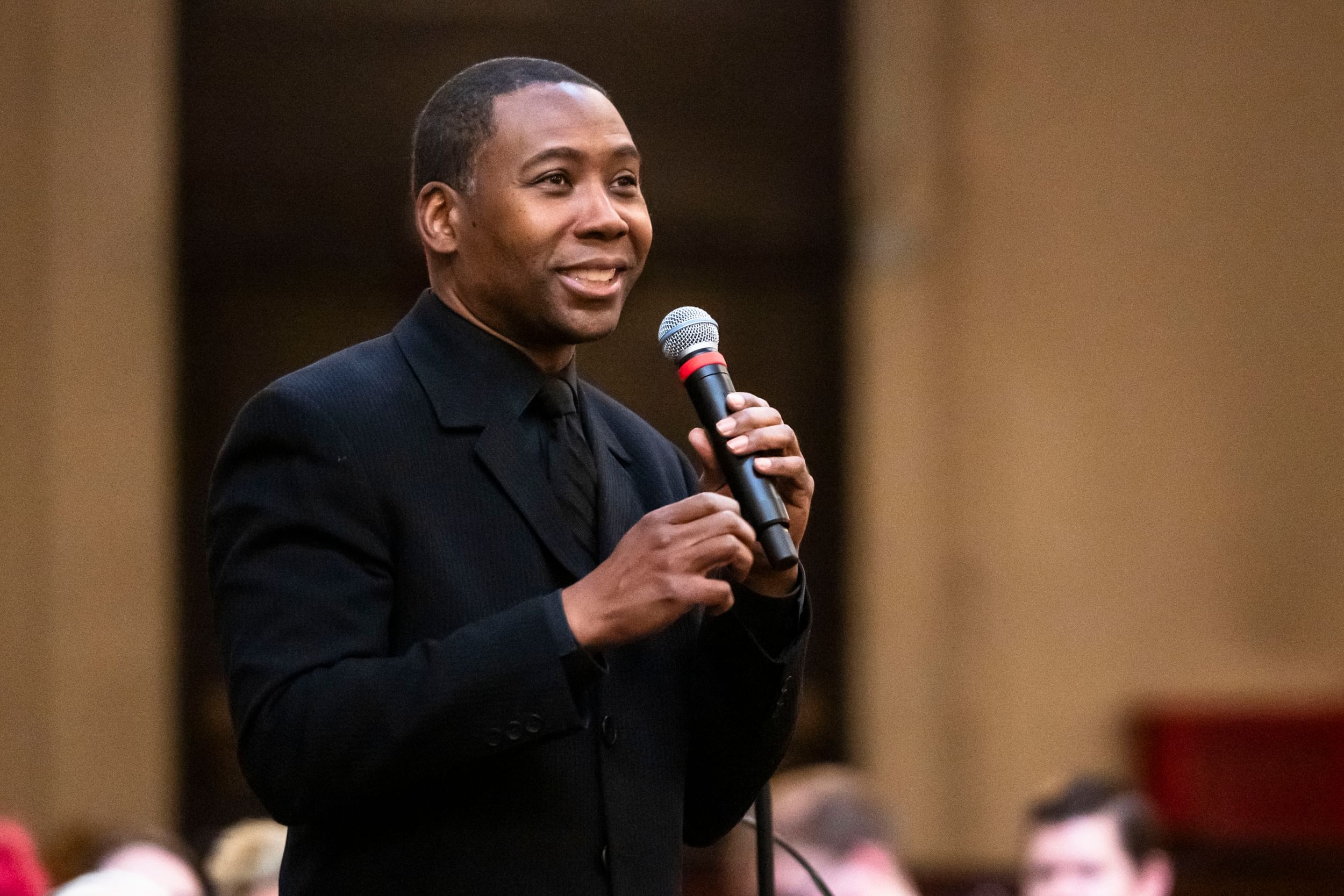 A Black man in an all-black suit on a stage, holding a microphone