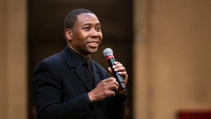 A Black man in an all-black suit on a stage, holding a microphone