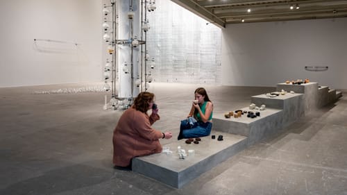 Wide gallery view with two people smelling handheld objects chosen from a variety on a low platform with graduated steps.