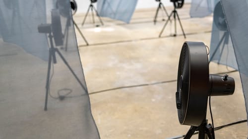 11 black fans on tripods point in different directions, blowing large, flowing transparent gray curtains with their breeze.