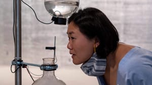 Seen in profile, an Asian woman wearing blue sniffs intently at a glass beaker mounted in the ICA gallery.