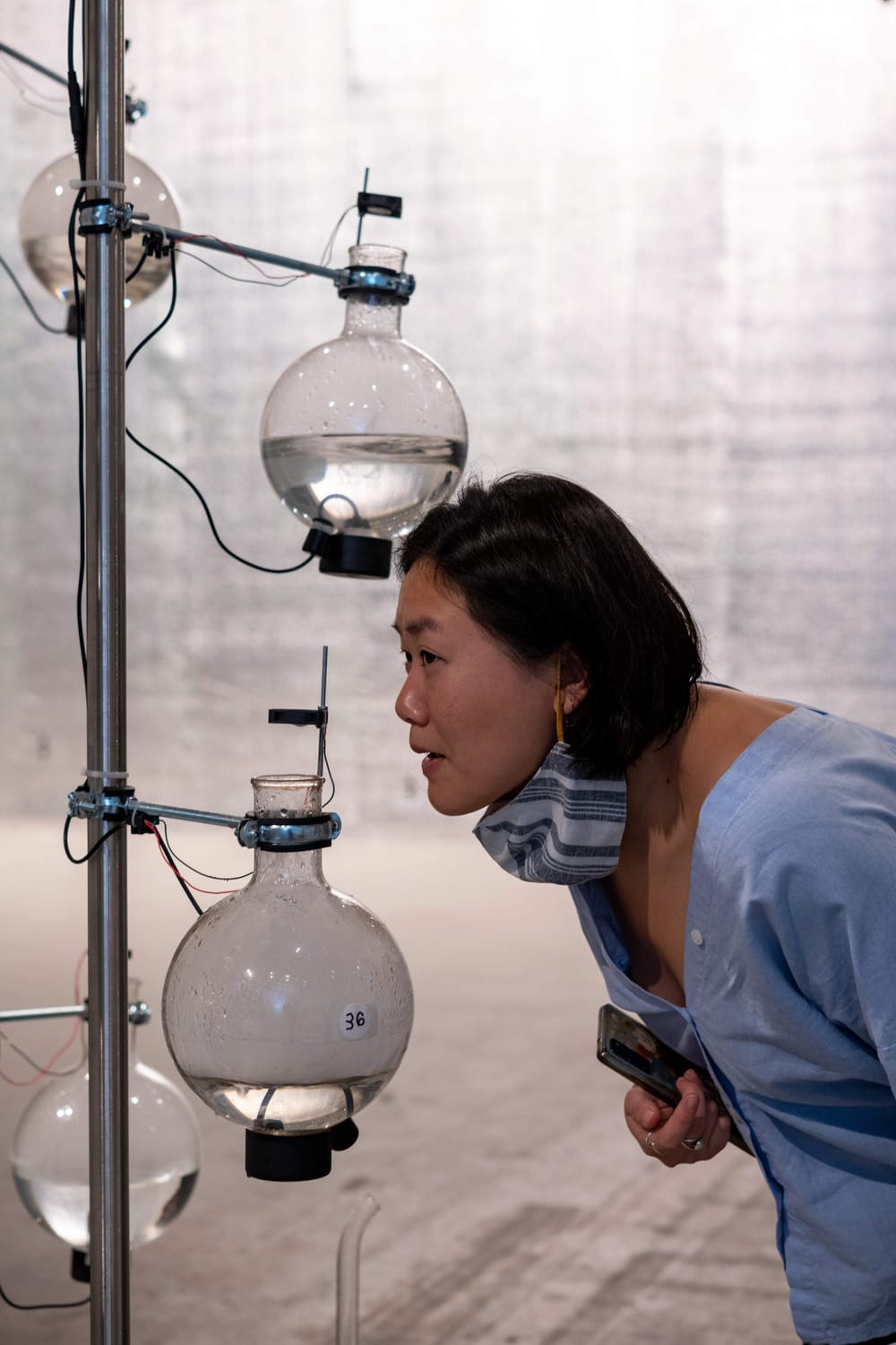 Seen in profile, an Asian woman wearing blue sniffs intently at a glass beaker mounted in the ICA gallery.