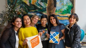 Six women pose for a group photo in front of a painting. Two hold books, one holds a Cookbooks & Convos tote