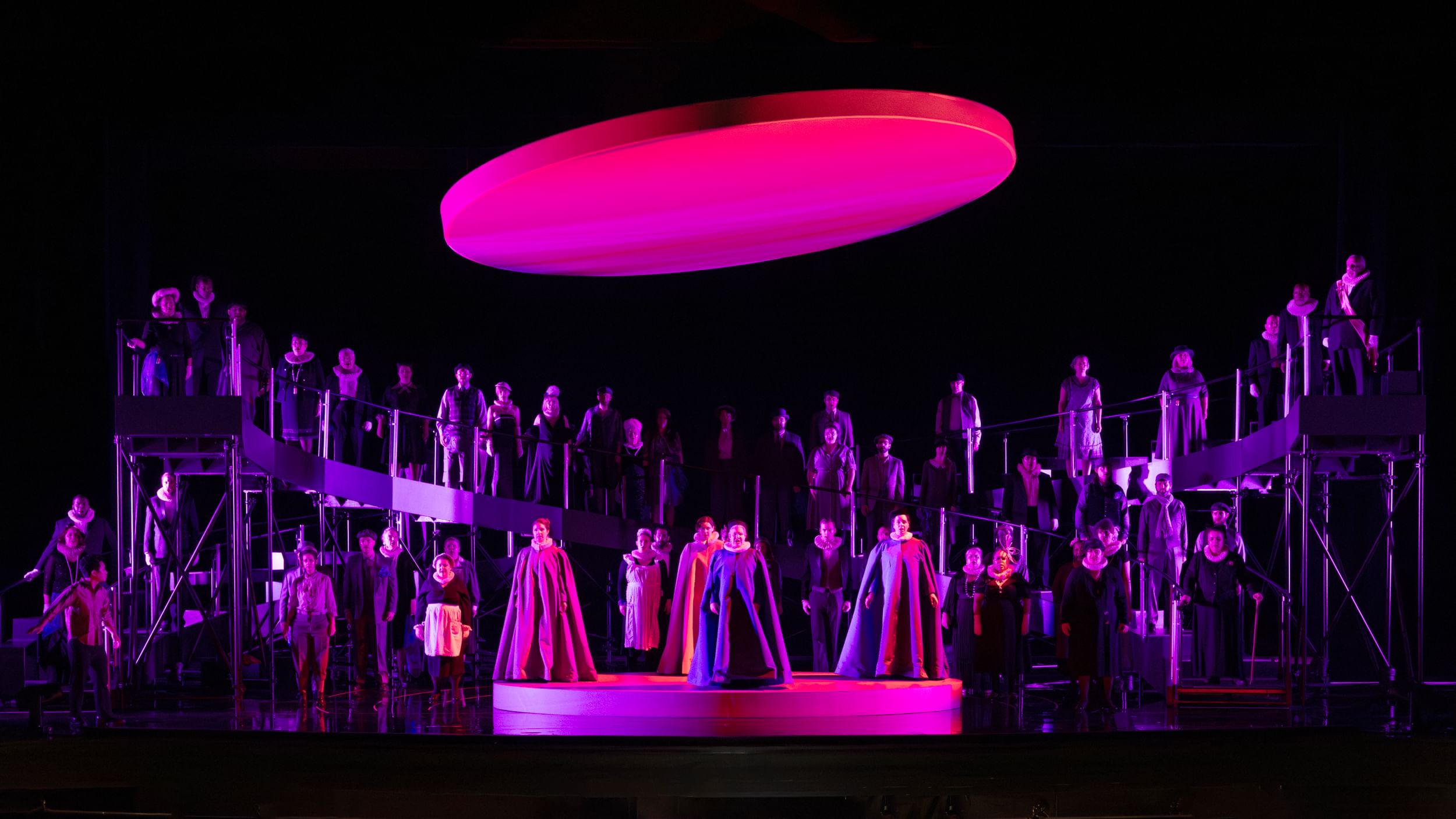 A few dozen chorus members surround four cloaked principles on a dais, under a large suspended disk, bathed in magenta light