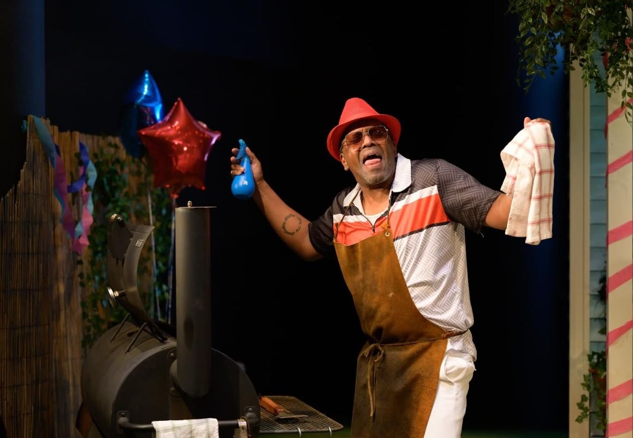 Smiling, in an apron and red fedora, stands exuberantly at an open grill, holding a rag and a spray bottle.