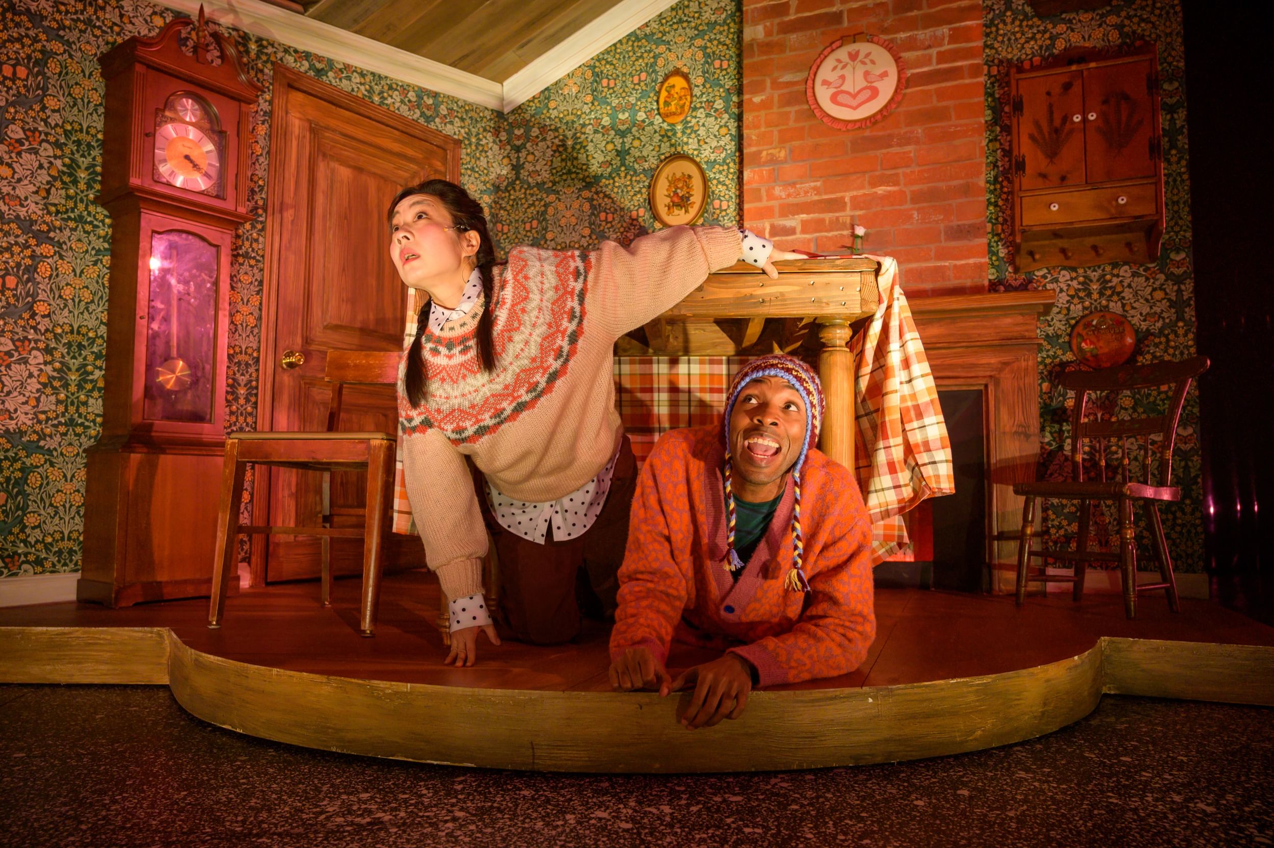 Ngo & Pierce, in cozy patterned sweaters, emerge surprised from under a table in a small floral, brick & plaid kitchen