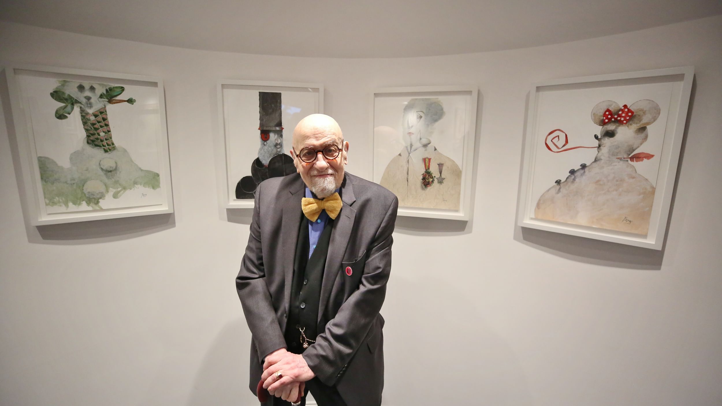 Paone, an elderly bald white man with round glasses & yellow bowtie, poses smiling in front of 4 snowpersons artworks.