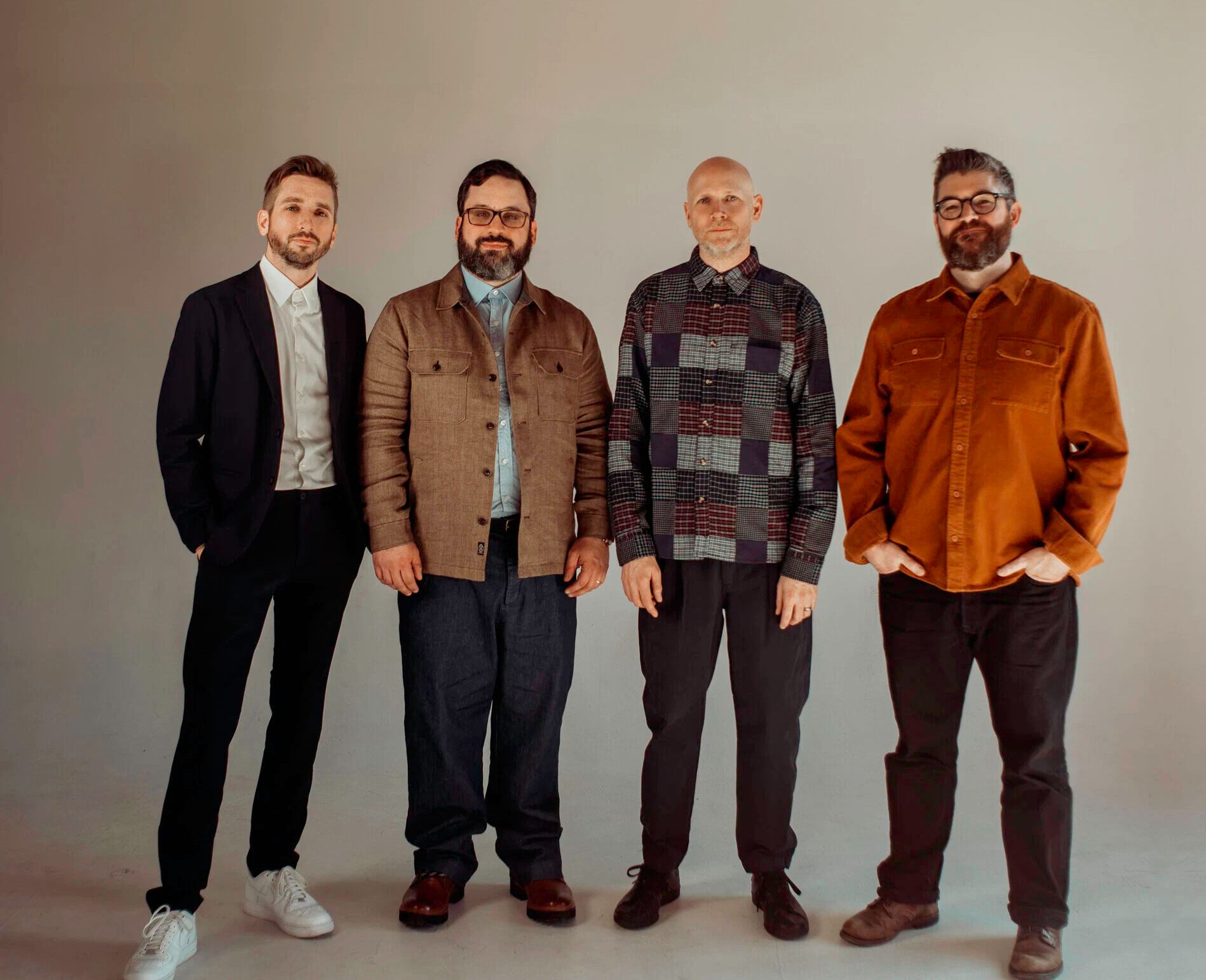 Group portrait. Four people stand next to each other, in jackets and button ups and solid pants, against a backdrop.