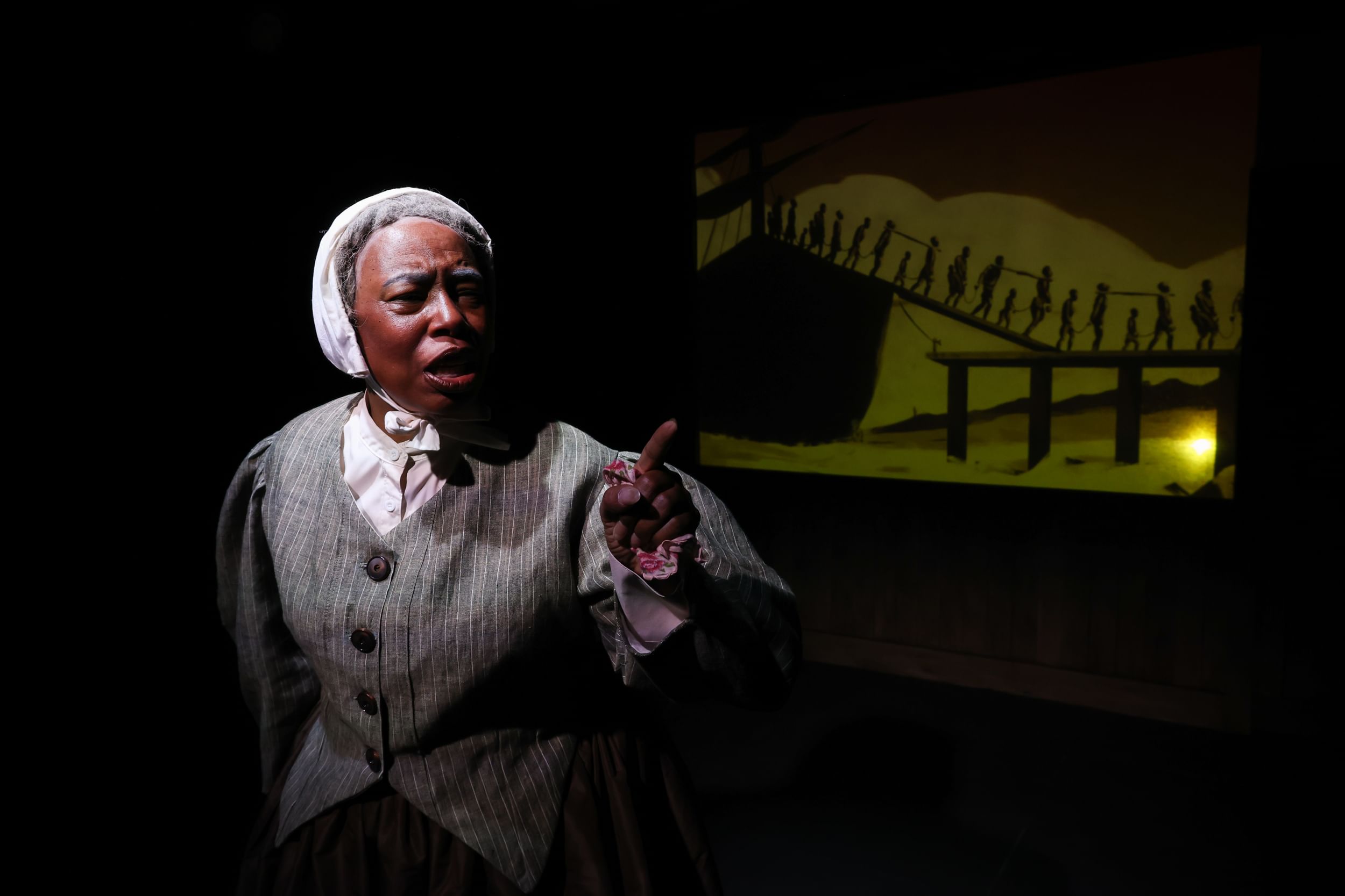 McGill, a Black woman in gray 19th-century costume, speaks onstage with a projection of enslaved people behind her.