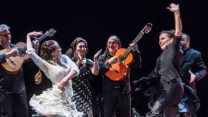 A pair of flamenco dancers in sevillana dresses perform with musicians behind them on stage.