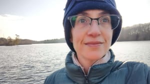 A selfie of Melissa, a white woman in her early 40s, by a pond. She wears a puffy coat, glasses, and a blue hat.