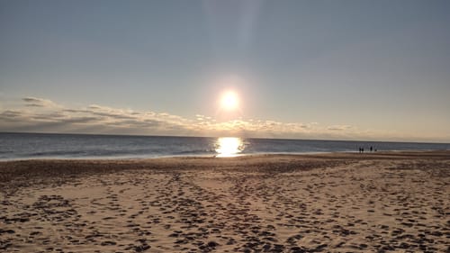 A photo of the ocean taken from the beach, the bright sun low over the shining water. The sand is covered with footprints.